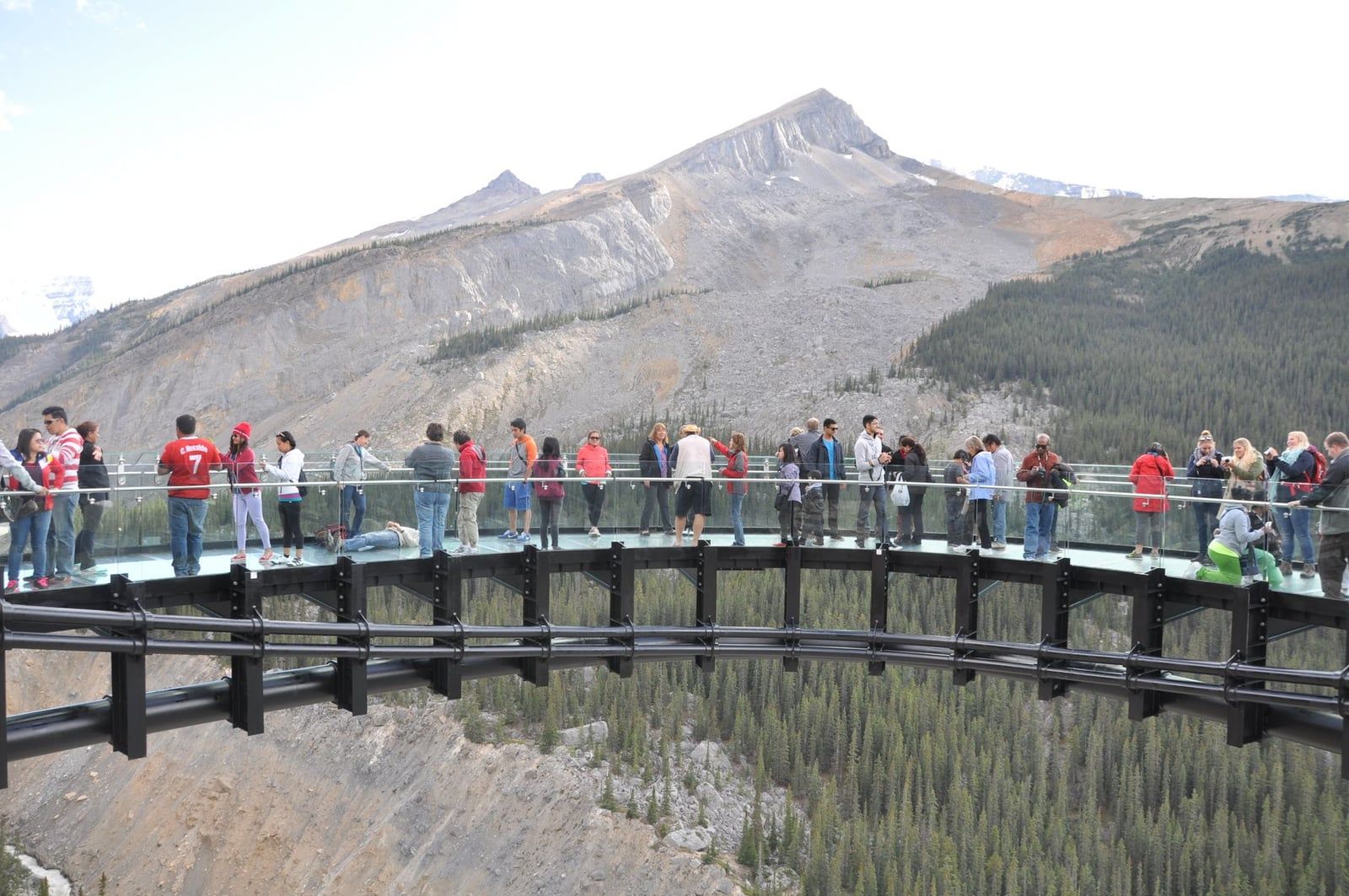 Visitors look out from the curved glass-floored Columbia Icefield Skywalk suspended high above a forested valley and rugged mountain peaks.